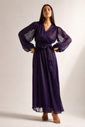 A woman modeling a long, dark purple pleated maxi dress with sheer balloon sleeves and a matching waist tie, standing against a plain light gray studio background.