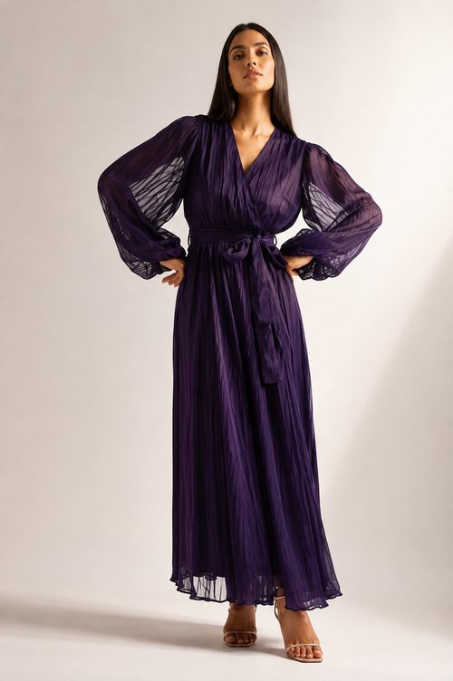 A woman modeling a long, dark purple pleated maxi dress with sheer balloon sleeves and a matching waist tie, standing against a plain light gray studio background.